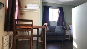 Une pièce chaleureuse avec une table et des chaises en bois, un canapé gris agrémenté de coussins à motifs, une petite table de chevet avec une lampe et une fenêtre avec des rideaux.