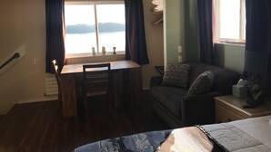 Une pièce comprenant un bureau en bois, une chaise, un canapé et un lit. Il y a une fenêtre avec des rideaux, un ventilateur de plafond et une vue sur une étendue d’eau.