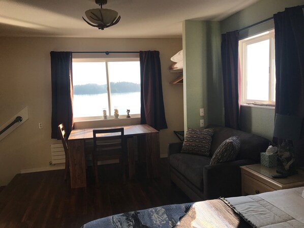 A room with a wooden desk, a chair, a sofa, and a bed. There is a window with curtains, a ceiling fan, and a view of a body of water. - Alaskan Waterfront Studio Apartment (Wolf Suite) Vacation Rental (Sitka)