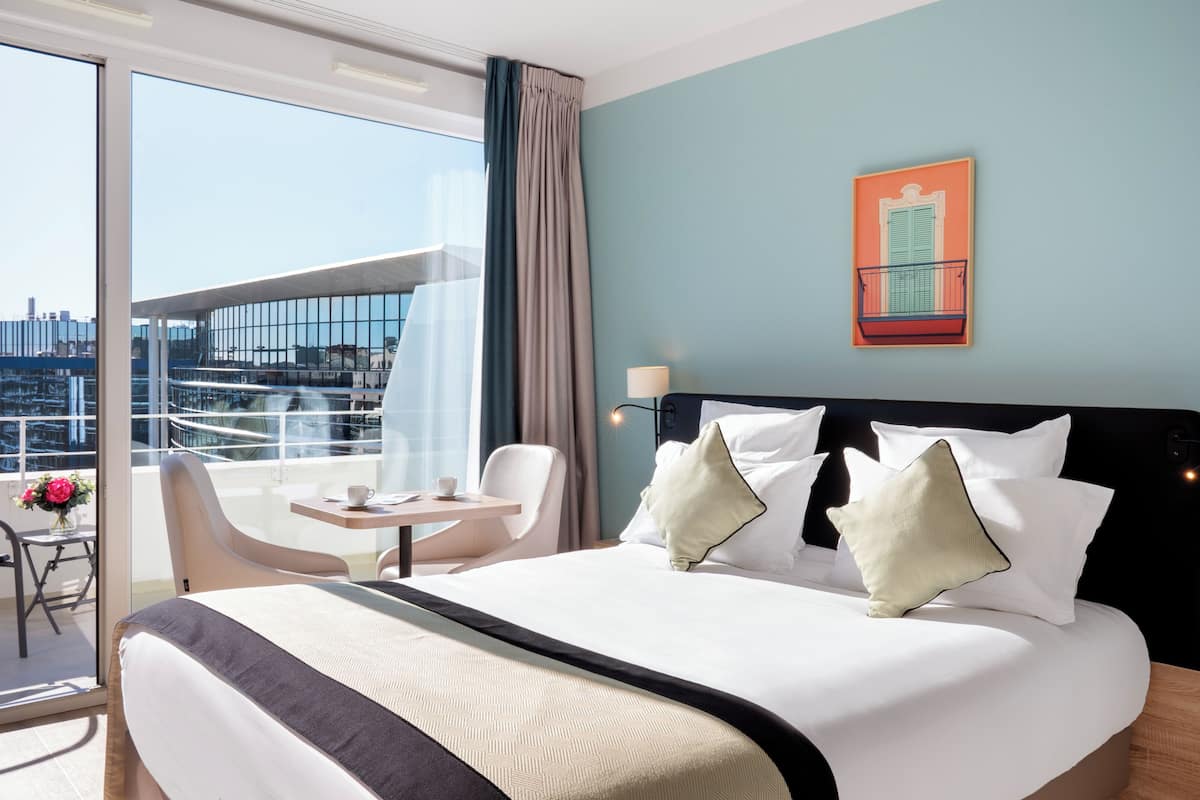 A hotel room with a large bed, a balcony with a chair and table, and a city view.