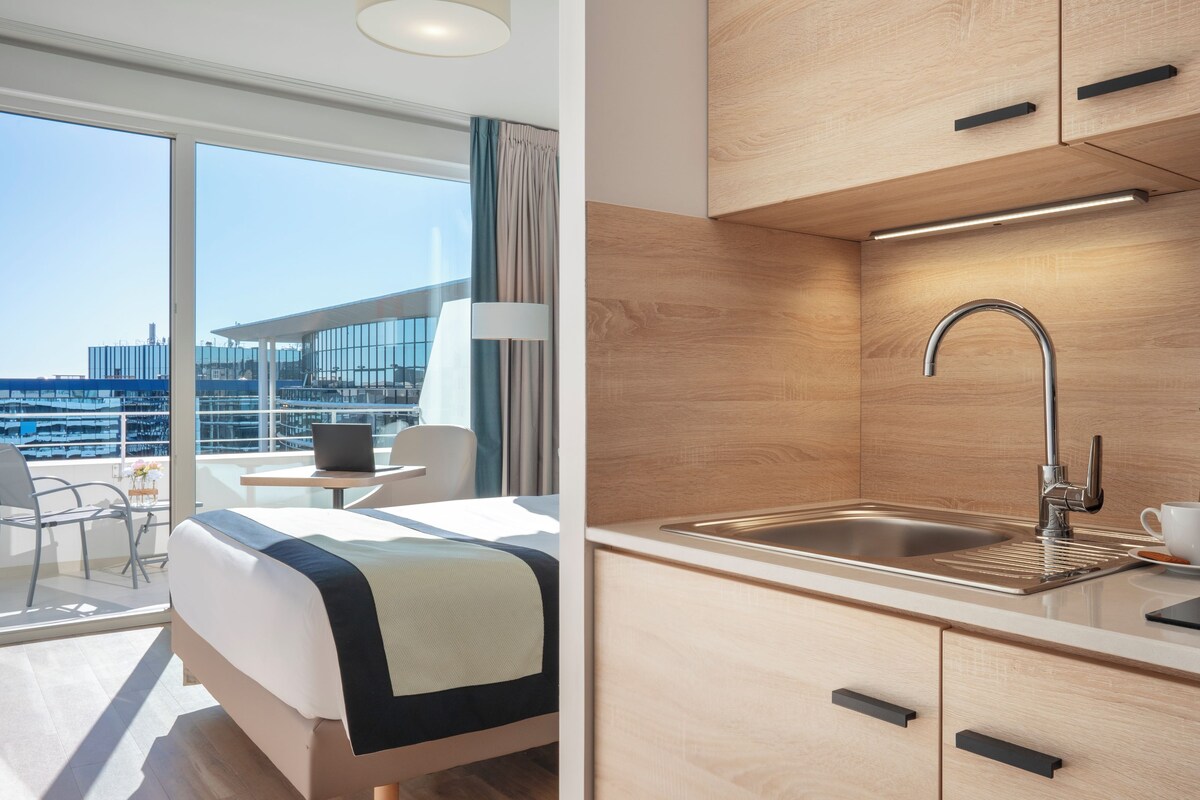 A modern hotel room with a balcony, a bed, a desk, and a kitchenette.