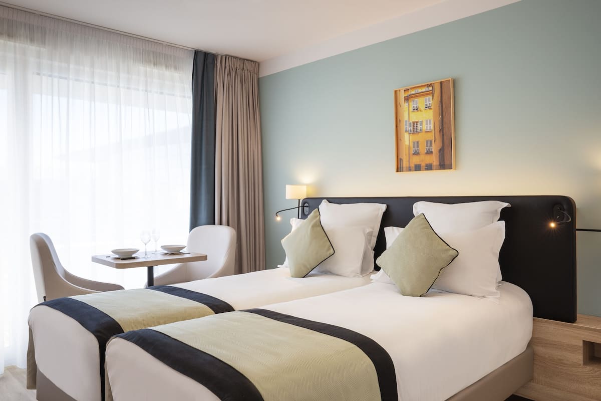 A hotel room with two beds, a small table with two wine glasses, a chair, and a painting on the wall.