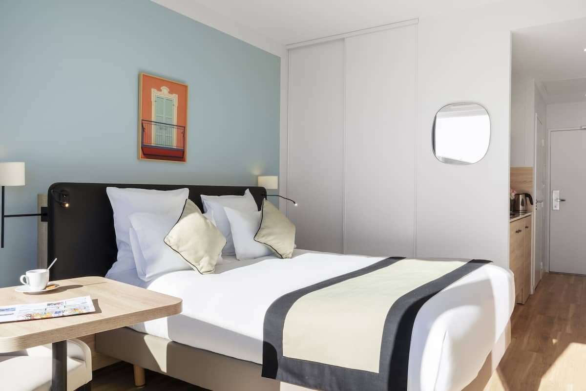 A hotel room with a bed, bedside tables, a chair, a small table with a cup and a menu, a painting on the wall, and a door leading to a bathroom.