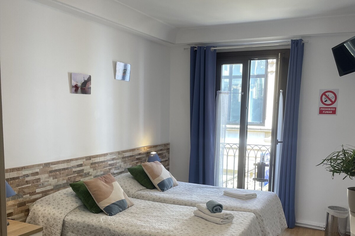 Hotellihuone, jossa on kaksi sänkyä, pieni työpöytä, roskakori ja näkymä ulos.