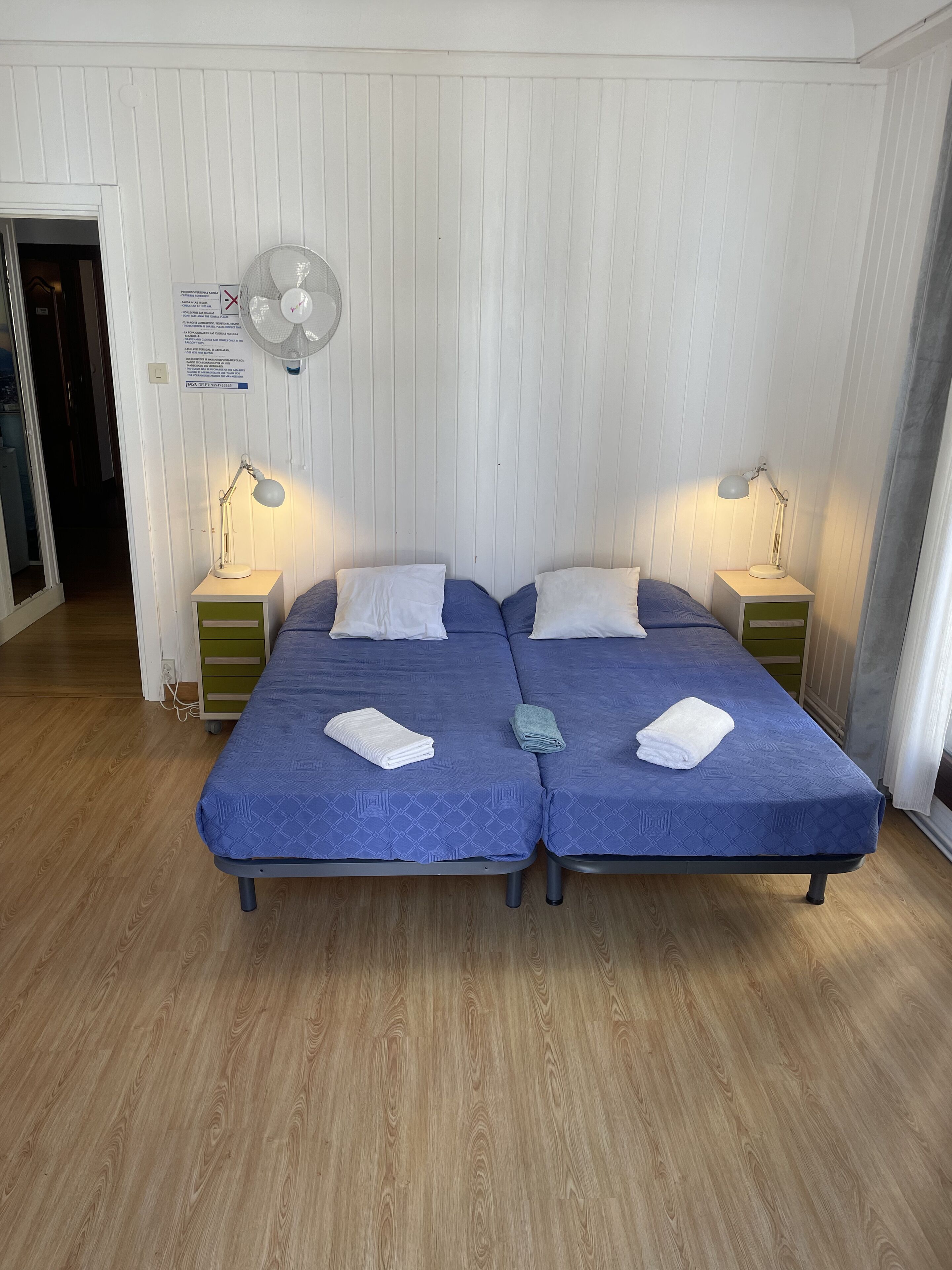 A room with two beds, each with a blue bedspread and white pillows, two bedside lamps, a wall-mounted fan, and a wooden floor.