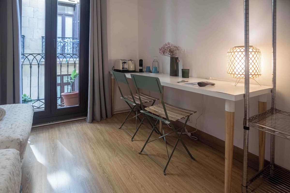 Habitación moderna con piso de madera, un escritorio blanco con lámpara, una silla y un balcón con vistas.