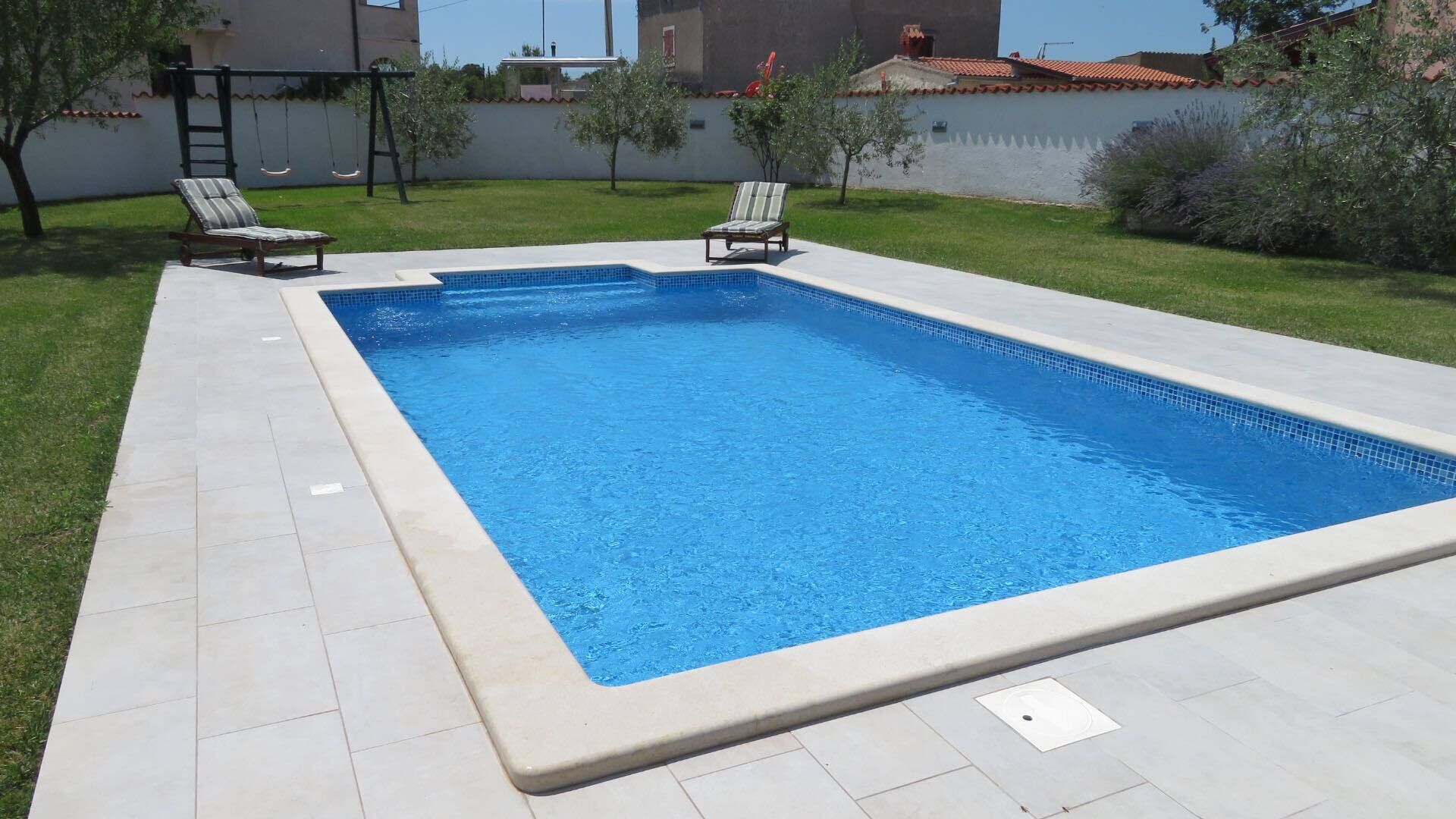 A rectangular swimming pool with a tiled edge, surrounded by a tiled deck. There are two lounge chairs on the deck.