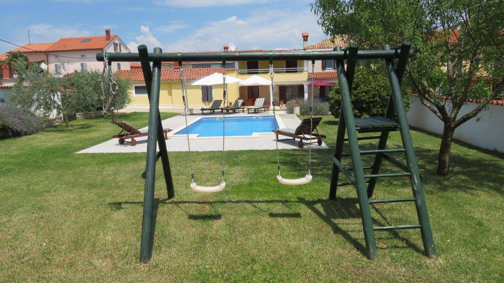 A backyard with a swing set, a pool, and a patio area with lounge chairs.