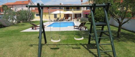 A backyard with a swing set, a pool, and a patio area with lounge chairs.