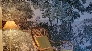 A wicker chair with a cushion in a room with a mural of trees and a lamp on a table.