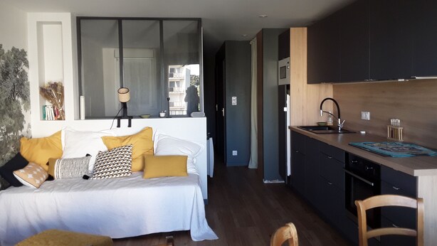 A modern bedroom with a large bed, bedside table, and a view into the adjacent kitchen area.