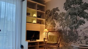 A living room with a wooden floor, a glass table, a wicker chair, a bookshelf, and a wall mural.