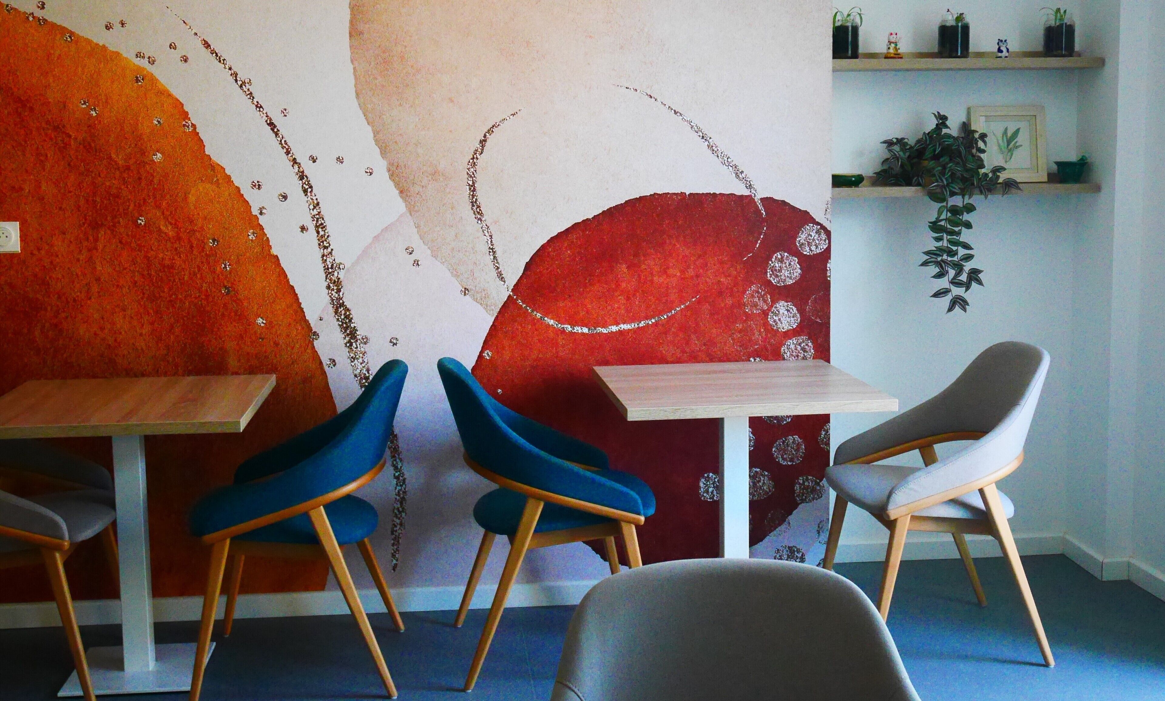 A dining area with two wooden tables and four chairs, one blue and one gray, against a wall with a large abstract painting.