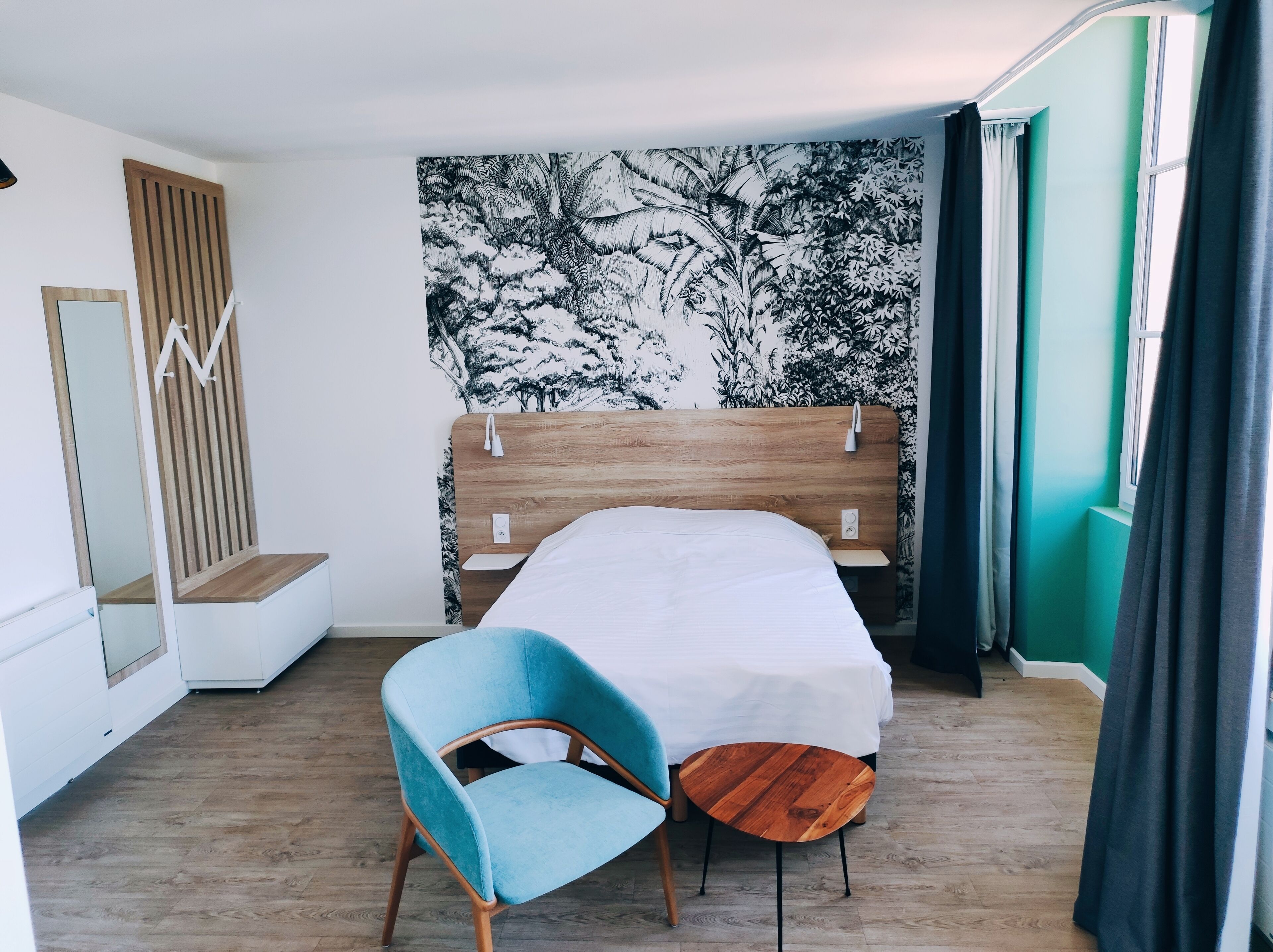 Une chambre avec un lit en bois, une chaise bleue, une petite table en bois, un miroir et un mur orné d’un motif d’arbre en noir et blanc.