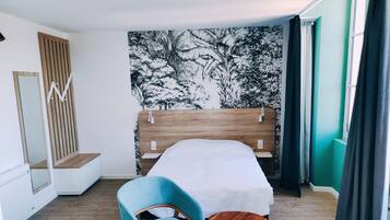 Une chambre avec un lit en bois, une chaise bleue, une petite table en bois, un miroir et un mur orné d’un motif d’arbre en noir et blanc.