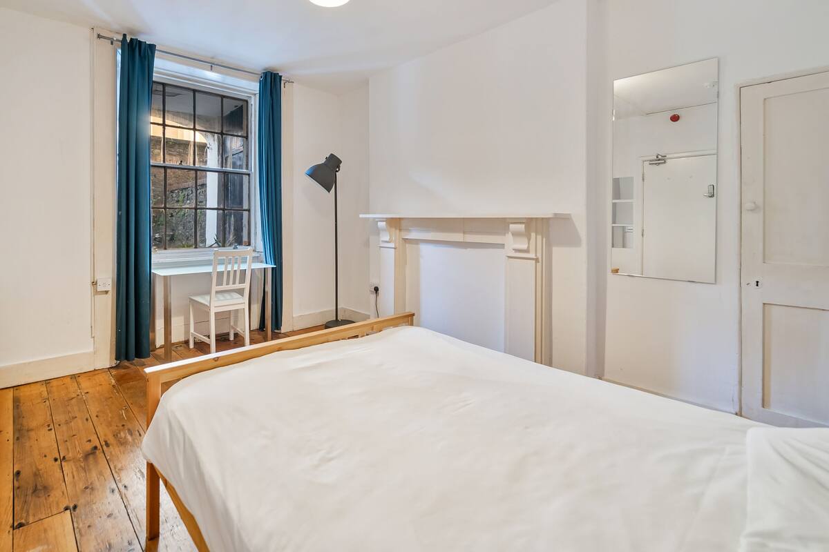 Une chambre avec un plancher en bois, un lit blanc, un bureau avec une chaise, une lampe et une fenêtre avec des rideaux.
