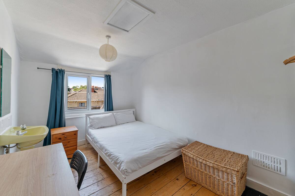 Une chambre à coucher comprenant un lit, une table de chevet, une chaise, une petite commode en bois, un panier en osier, une fenêtre avec des rideaux et un luminaire au plafond.
