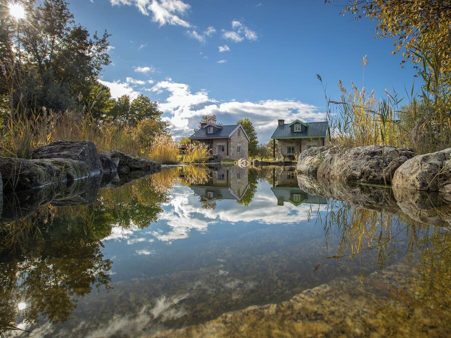 A serene lakeside scene with two stone houses reflecting in the water, surrounded by natural vegetation and a clear sky.