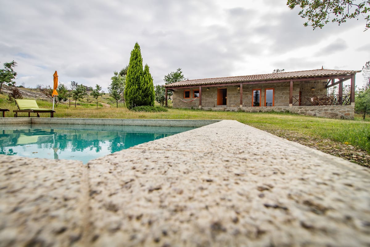 A stone house with a pool, a lounge chair, and a tall tree.