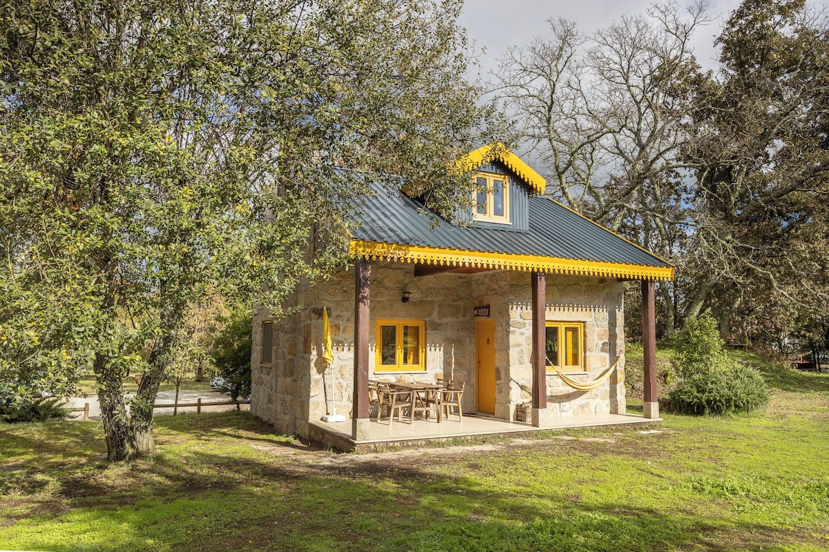 A stone cottage with a yellow roof and wooden porch, surrounded by trees.