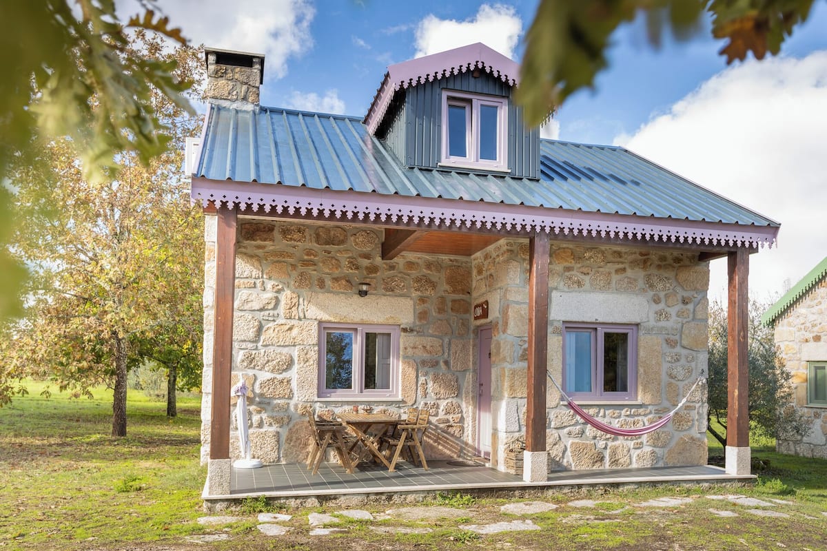 A stone house with a metal roof, two windows, and a wooden table outside.