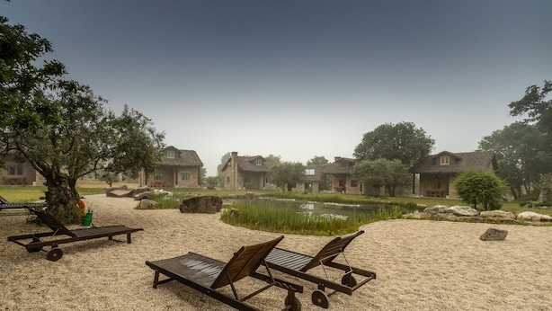 A serene outdoor area with thatched-roof cottages, a pond, and lounge chairs on a gravel surface. - Chão do Rio - Turismo de Aldeia (Seia)
