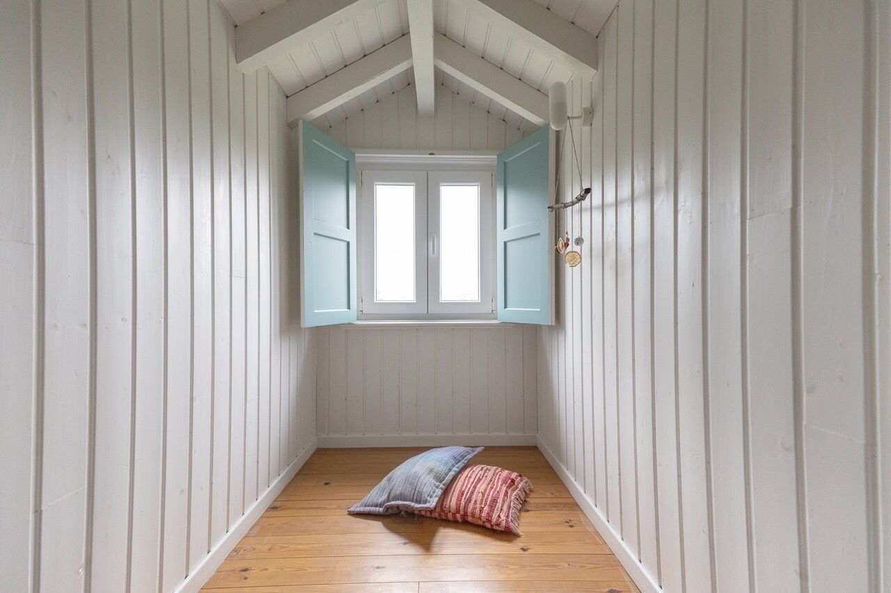 A narrow room with white wooden walls, a wooden floor, and a window with blue shutters.
