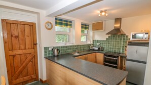 A kitchen with a wooden door, a clock, a window with blinds, a microwave, and a refrigerator. - Garreg Lwyd Farm (Pwllheli)