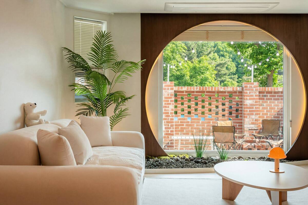 A modern living room with a sofa, a round table, and a large window with a view of a brick wall and greenery.