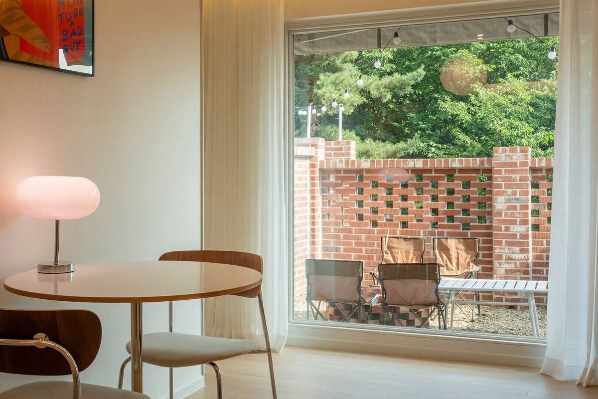 A dining area with a table and chairs, a lamp, and a view of a brick wall and greenery outside.