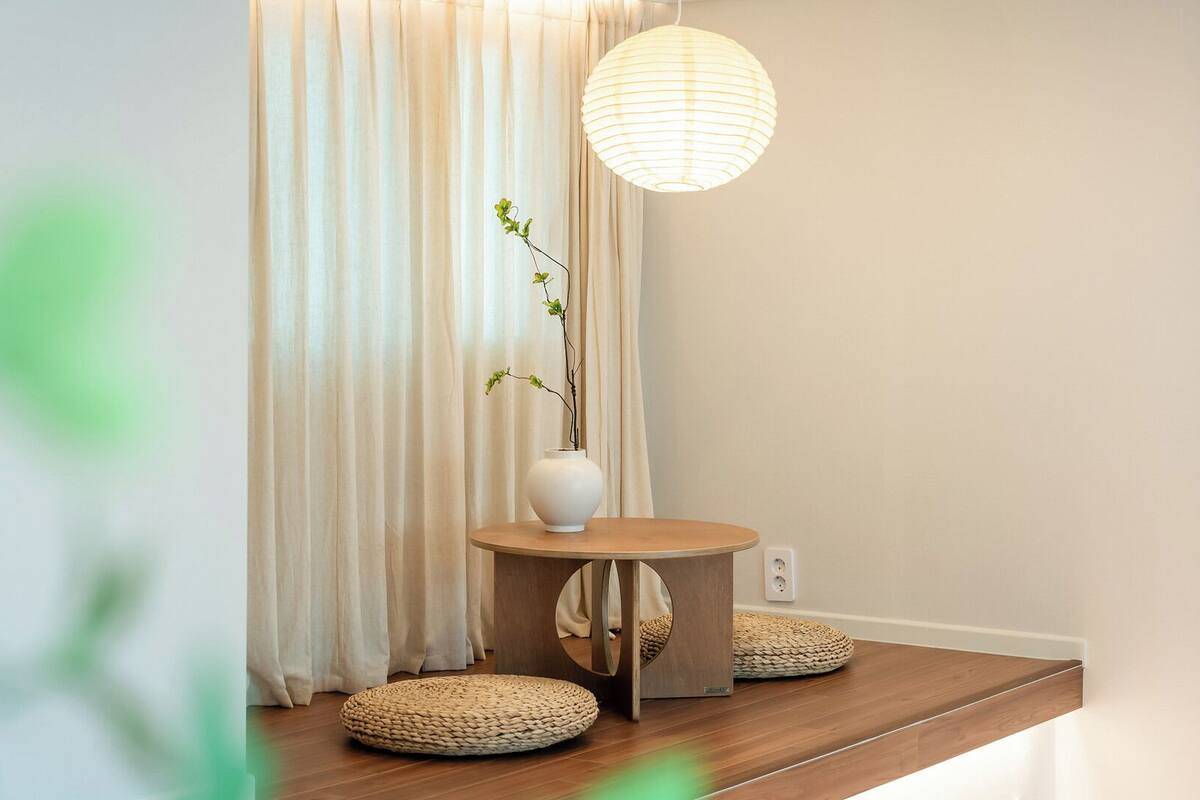A room with a round wooden table, a white vase with a plant, a woven pouf, and a hanging paper lantern.