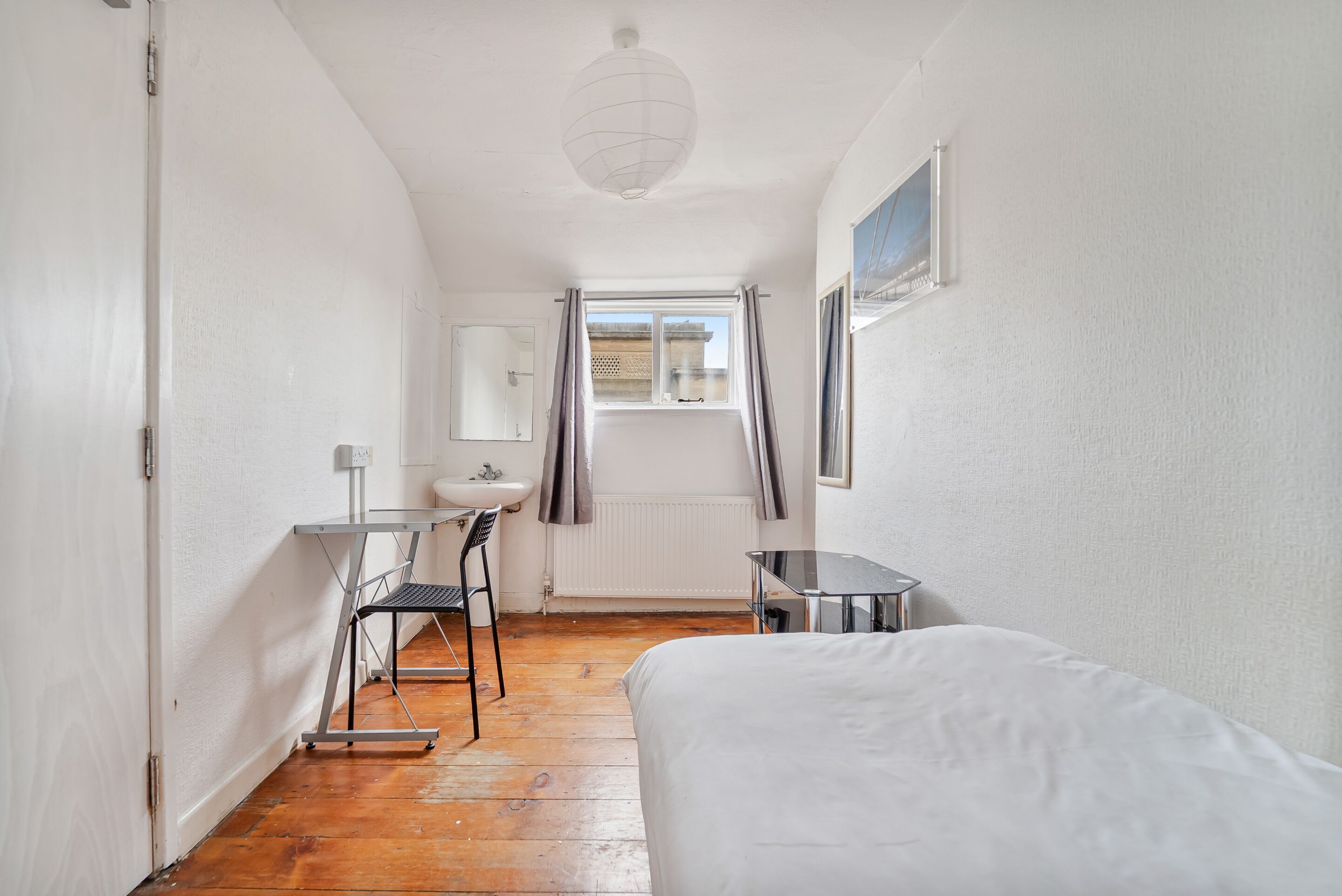 Une petite pièce lumineuse, avec un lit, un bureau et une chaise. Il y a une fenêtre avec des rideaux, un radiateur et un tableau accroché au mur.