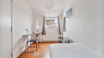 Une petite pièce lumineuse, avec un lit, un bureau et une chaise. Il y a une fenêtre avec des rideaux, un radiateur et un tableau accroché au mur.