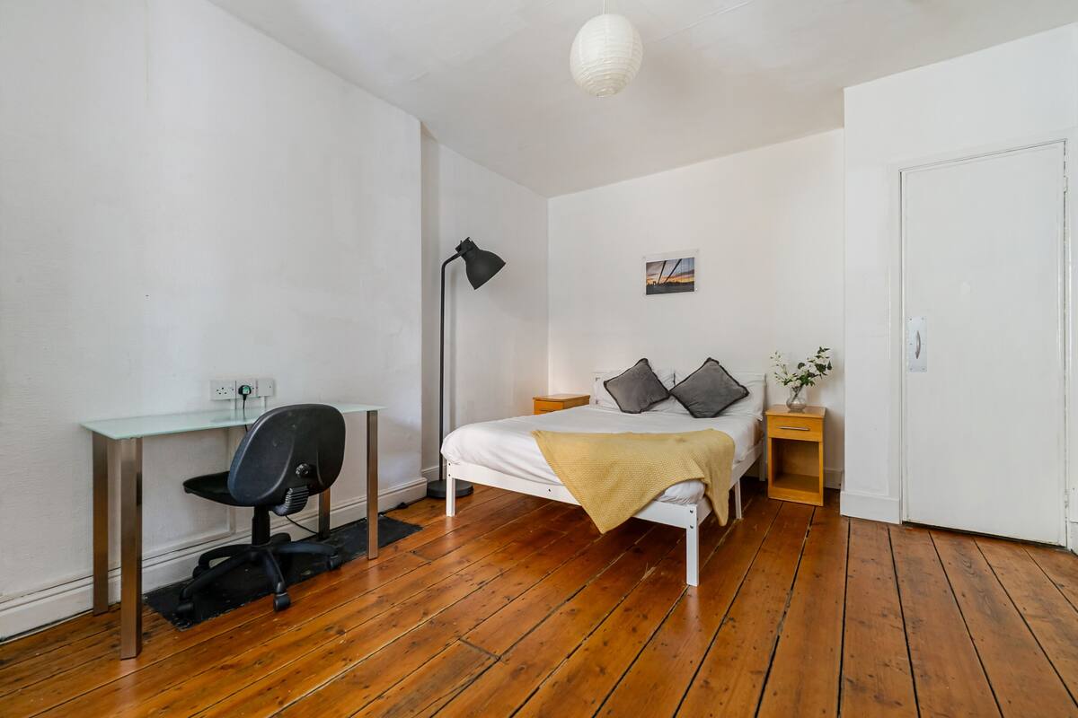 Un dormitorio con piso de madera, una cama con almohadas, un escritorio con una silla, una lámpara y una mesita de noche con una planta.