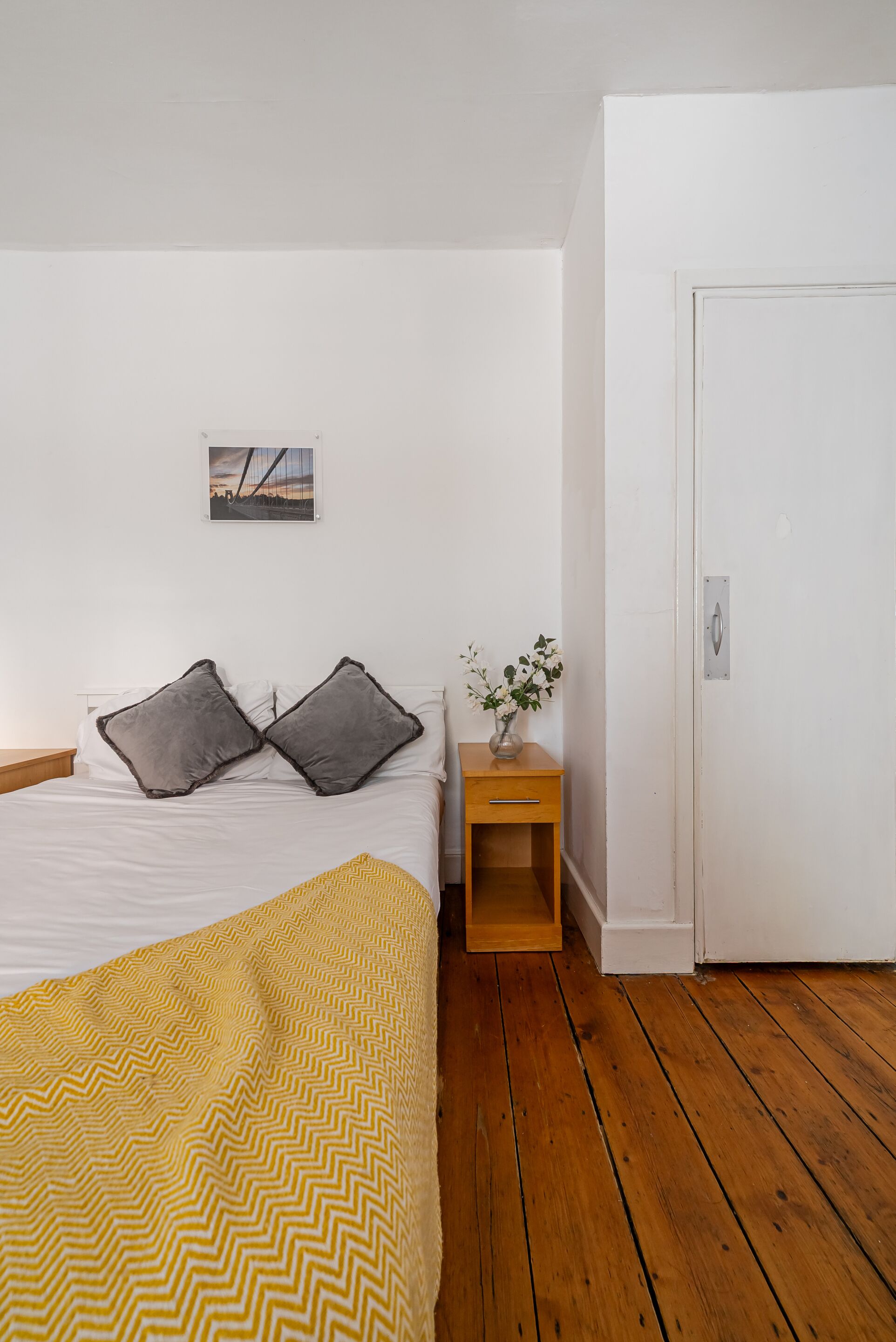 A bedroom with a bed, two pillows, a nightstand with a vase, and a door.