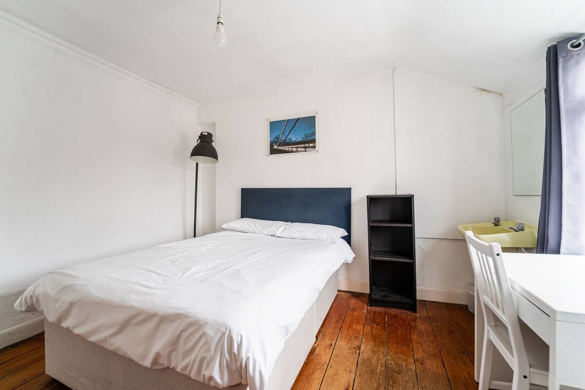 Un dormitorio con cama, escritorio, silla, estante y un cuadro en la pared.