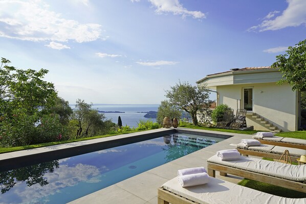 A swimming pool with lounge chairs and towels, surrounded by greenery and a view of the sea.