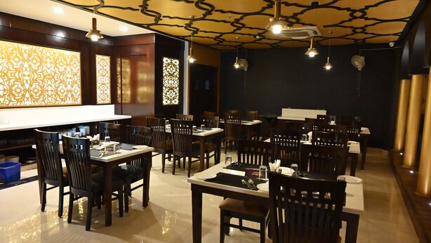 A restaurant with multiple tables and chairs, a patterned ceiling, and decorative wall panels. - The Triumph Hotel (Varanasi)