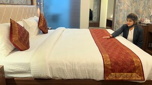A neatly made bed with a red and gold bedspread, a white pillow, and a decorative pillow. - The Triumph Hotel (Varanasi)