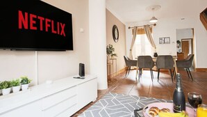 Ein modernes Wohnzimmer mit einem Flachbildfernseher, der das Netflix-Logo zeigt, einem Essbereich mit Tisch und Stühlen sowie einer Küchenzeile mit Spüle und Arbeitsplatte.