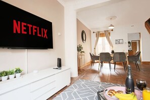 A modern living room with a flat-screen TV displaying the Netflix logo, a dining area with a table and chairs, and a kitchen area with a sink and countertop.