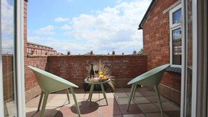 Eine Terrasse mit zwei grünen Stühlen, einem kleinen runden Tisch mit einer Flasche und Obst, und einer Backsteinmauer.