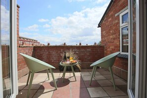 A patio with two green chairs, a small round table with a bottle and fruit, and a brick wall.