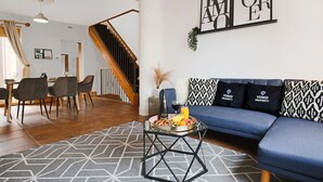 A modern living room with a blue sofa, a dining area with a wooden table and chairs, and a staircase.