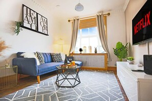A living room with a blue sofa, a coffee table with fruits, a TV, and a window with curtains. - Central House • Sleeps 9 • FreeParking • Pool Table • Fast Wifi • Balcony • VirginTV • Yoko Property (Northampton)