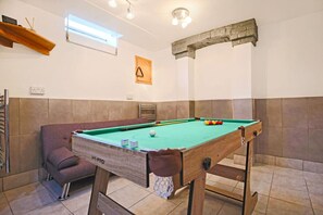 A pool table in a room with a brown sofa, a wooden shelf, and a window.
