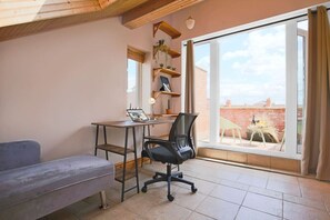 A small office space with a desk, chair, and a sofa. There is a window with a view of a brick building outside.