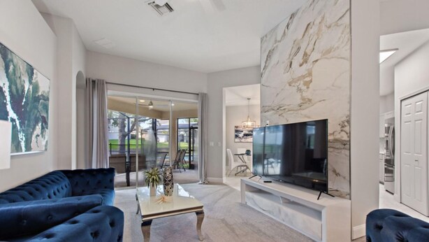 A modern living room with a large flat-screen TV, plush blue sofas, a marble accent wall, and a sliding glass door leading to an outdoor area.