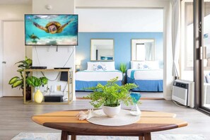 A modern hotel room with a large TV, two beds, a dining table, and a potted plant.