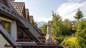 A residential building with a sloped roof, wooden balcony railings, and a view of a lush green landscape with trees and mountains in the background.